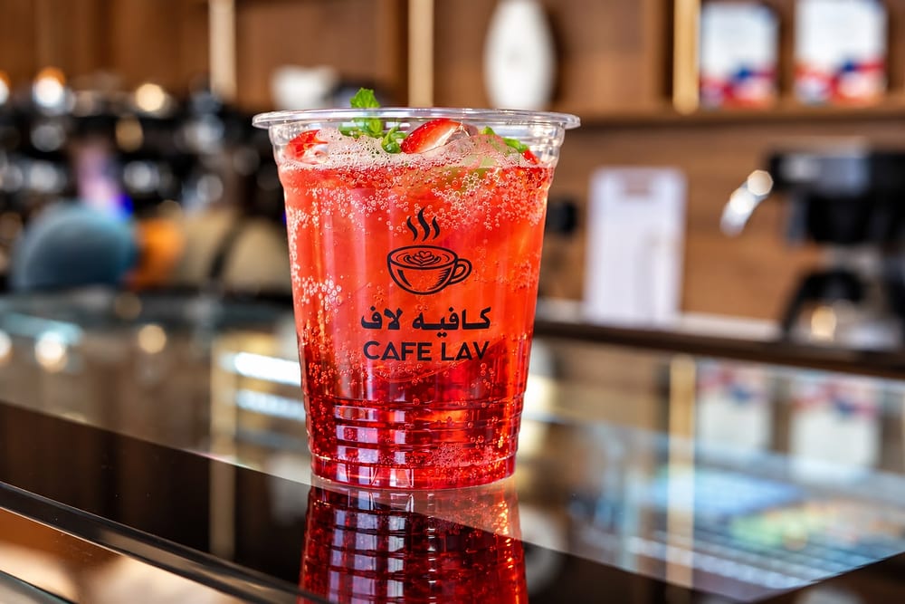 كوب مشروب أحمر مثلّج بالفراولة والنعناع يحمل شعار كافيه لاف CAFE LAV على منضدة زجاجية، وخلفه آلة القهوة والرفوف